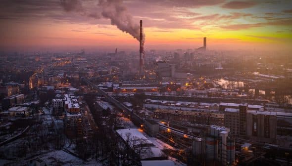 Aerial view of an industrial area with smoke stacks emitting smoke against a sunset sky. - Olive Oil Times