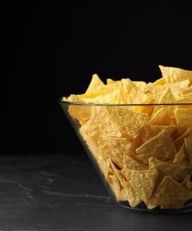 A clear glass bowl filled with yellow tortilla chips on a dark surface. - Olive Oil Times