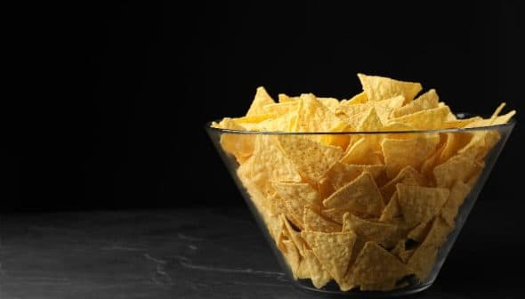 A clear glass bowl filled with yellow tortilla chips on a dark surface. - Olive Oil Times
