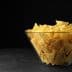 A clear glass bowl filled with yellow tortilla chips on a dark surface. - Olive Oil Times