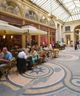 People dining at tables in a covered arcade with a glass roof and decorative floor tiles. - Olive Oil Times