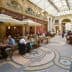 People dining at tables in a covered arcade with a glass roof and decorative floor tiles. - Olive Oil Times