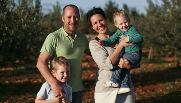 A family of four posing together in an olive grove, with two children and two adults smiling at the camera. - Olive Oil Times