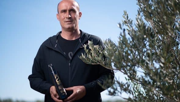 Man standing outdoors holding a bottle of olive oil next to an olive tree. - Olive Oil Times