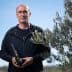 Man standing outdoors holding a bottle of olive oil next to an olive tree. - Olive Oil Times