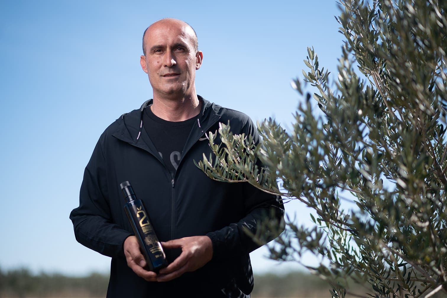 Man standing outdoors holding a bottle of olive oil next to an olive tree. - Olive Oil Times