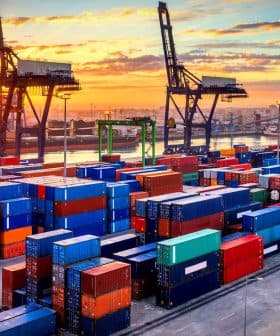Colorful shipping containers stacked at a port with cranes in the background during sunset. - Olive Oil Times