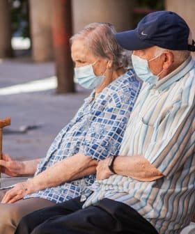An elderly couple wearing masks sitting on a bench, with one holding a cane. - Olive Oil Times