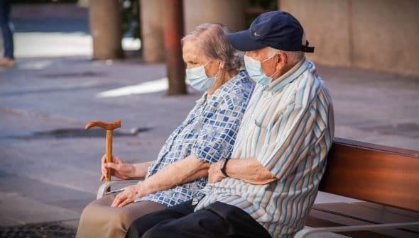 An elderly couple wearing masks sitting on a bench, with one holding a cane. - Olive Oil Times