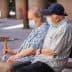 An elderly couple wearing masks sitting on a bench, with one holding a cane. - Olive Oil Times