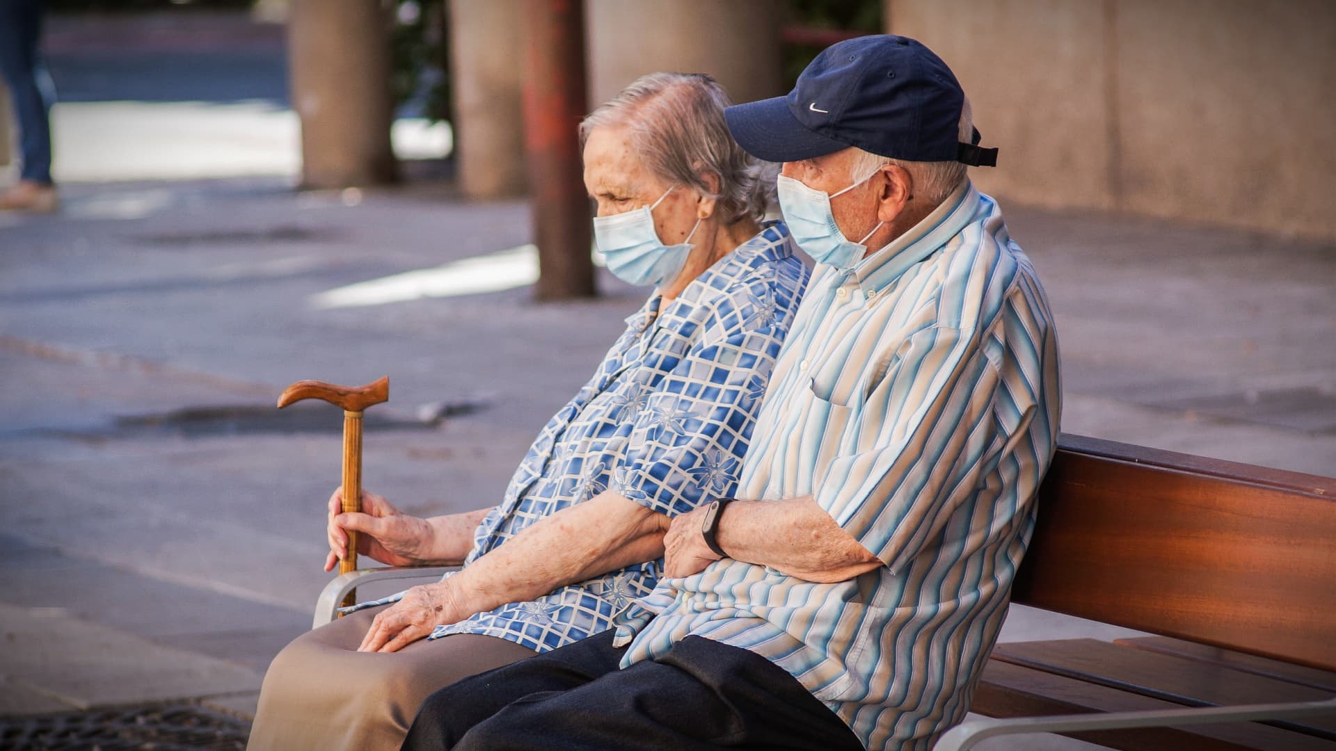An elderly couple wearing masks sitting on a bench, with one holding a cane. - Olive Oil Times