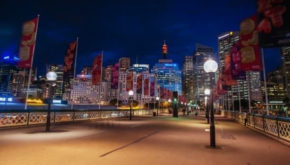 Night view of Sydney cityscape featuring illuminated buildings and decorative flags along a walkway. - Olive Oil Times