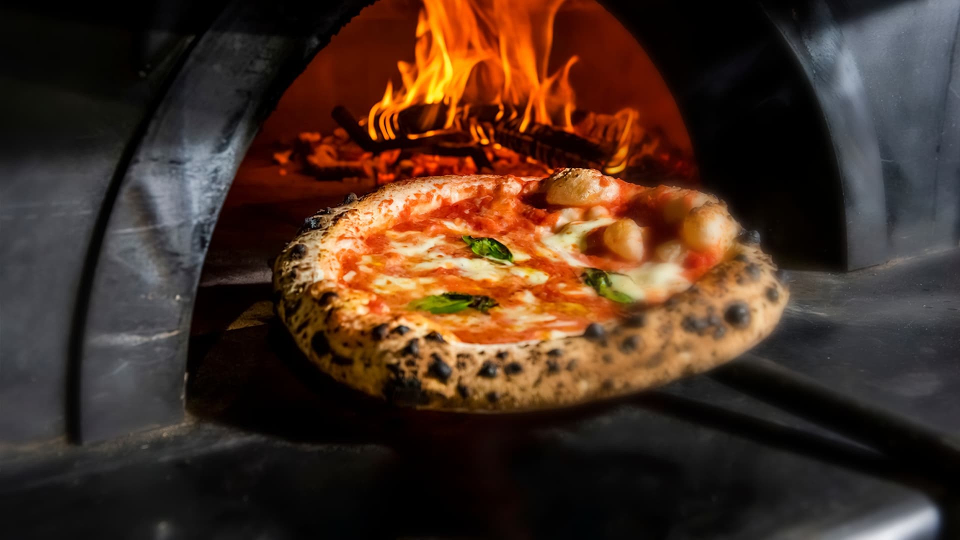 A freshly baked pizza with melted cheese and basil being pulled from a wood-fired oven. - Olive Oil Times