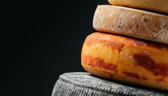 A stack of different types of cheese, including a yellow-orange cheese and a dark cheese, against a dark background. - Olive Oil Times