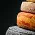 A stack of different types of cheese, including a yellow-orange cheese and a dark cheese, against a dark background. - Olive Oil Times