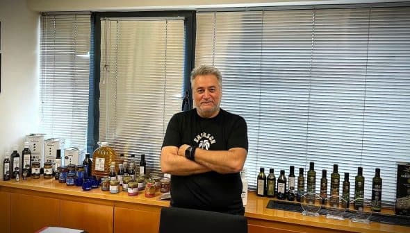 Man with gray hair wearing a black t-shirt standing in an office with olive oil products displayed on a shelf. - Olive Oil Times