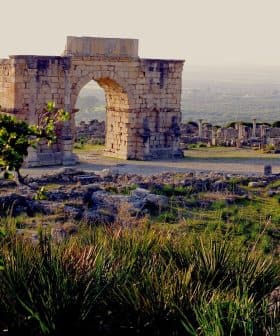 Stone archway structure at an archaeological site with surrounding ruins and greenery. - Olive Oil Times