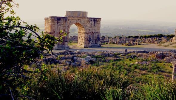 Stone archway structure at an archaeological site with surrounding ruins and greenery. - Olive Oil Times