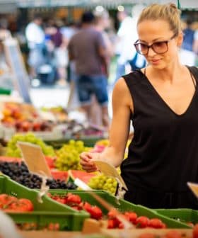 A woman wearing sunglasses examines produce at a farmers market with various fruits and vegetables displayed. - Olive Oil Times