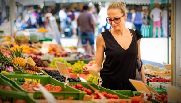 A woman wearing sunglasses examines produce at a farmers market with various fruits and vegetables displayed. - Olive Oil Times