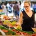 A woman wearing sunglasses examines produce at a farmers market with various fruits and vegetables displayed. - Olive Oil Times