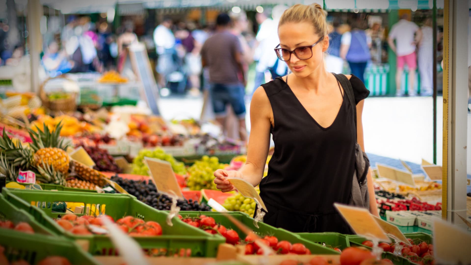 A woman wearing sunglasses examines produce at a farmers market with various fruits and vegetables displayed. - Olive Oil Times