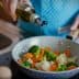 A person in an apron drizzling olive oil over a pan of mixed vegetables including broccoli and carrots. - Olive Oil Times