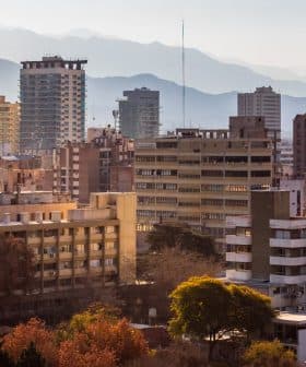 A cityscape featuring various buildings with mountains visible in the background under a clear sky. - Olive Oil Times