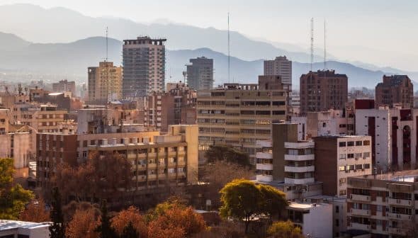 A cityscape featuring various buildings with mountains visible in the background under a clear sky. - Olive Oil Times