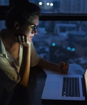 Woman with glasses writing notes while looking at a laptop in a dimly lit environment at night. - Olive Oil Times