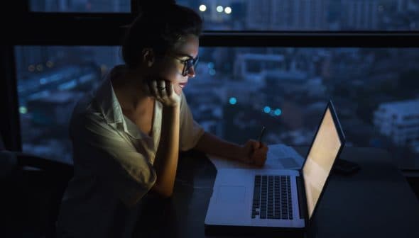 Woman with glasses writing notes while looking at a laptop in a dimly lit environment at night. - Olive Oil Times