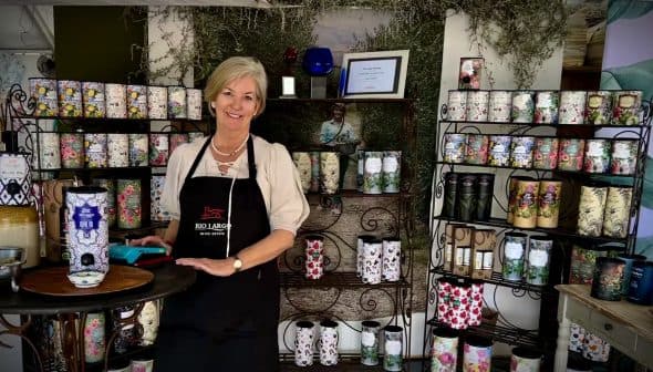 A woman wearing an apron stands beside a display of decorative products on shelves. - Olive Oil Times