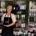 A woman wearing an apron stands beside a display of decorative products on shelves. - Olive Oil Times