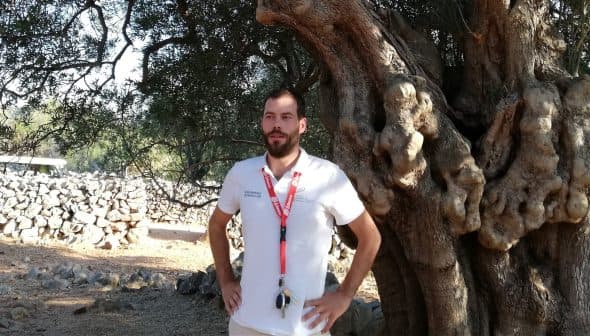 A man wearing a white shirt and lanyard standing beside a large olive tree. - Olive Oil Times