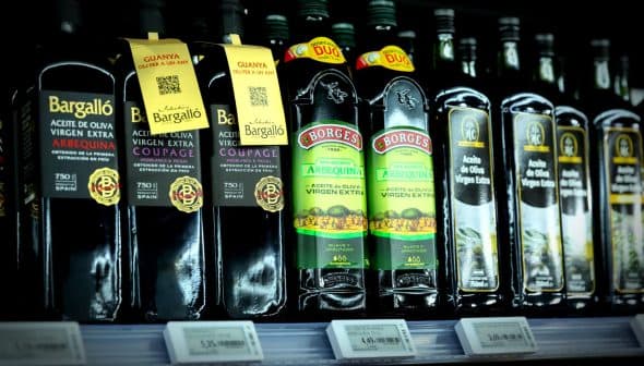 Various bottles of olive oil displayed on a store shelf with labels and pricing information. - Olive Oil Times