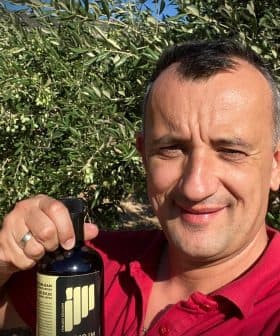 Man smiling while holding a bottle of olive oil in an olive grove. - Olive Oil Times