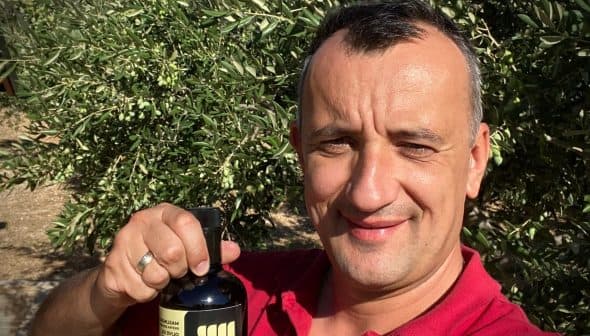 Man smiling while holding a bottle of olive oil in an olive grove. - Olive Oil Times