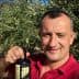 Man smiling while holding a bottle of olive oil in an olive grove. - Olive Oil Times