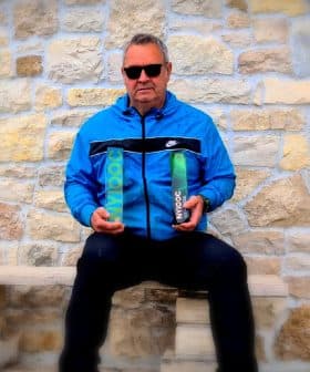 Man wearing sunglasses and a blue jacket holding two bottles of olive oil while seated on a stone bench. - Olive Oil Times
