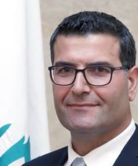 Man wearing glasses and a suit, standing in front of a Lebanese flag. - Olive Oil Times