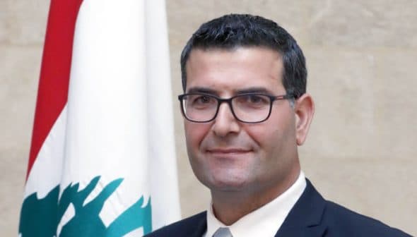 Man wearing glasses and a suit, standing in front of a Lebanese flag. - Olive Oil Times