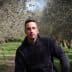A man wearing a black hoodie sitting among olive trees in an olive grove. - Olive Oil Times