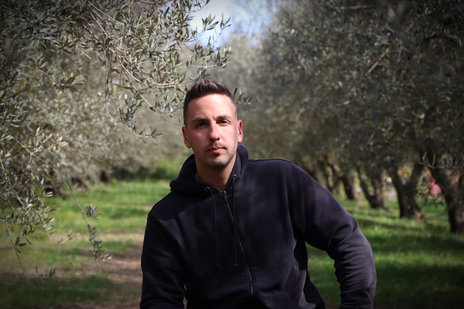 A man wearing a black hoodie sitting among olive trees in an olive grove. - Olive Oil Times