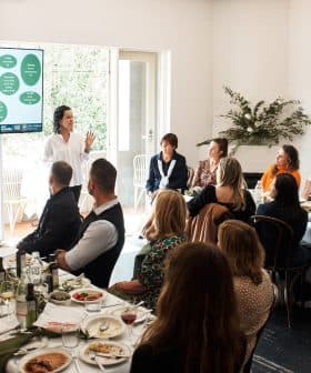 A speaker presenting information on a screen to an audience seated at a long table during a meeting. - Olive Oil Times