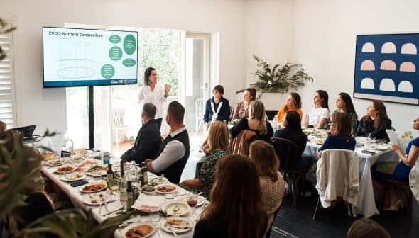 A speaker presenting information on a screen to an audience seated at a long table during a meeting. - Olive Oil Times