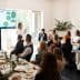 A speaker presenting information on a screen to an audience seated at a long table during a meeting. - Olive Oil Times