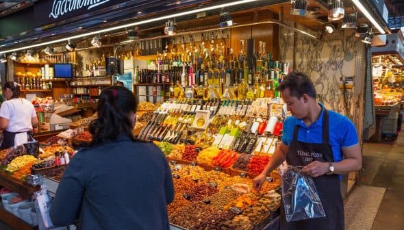 A market stall displaying various dried fruits, nuts, and bottles of olive oil with a vendor assisting a customer. - Olive Oil Times