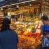 A market stall displaying various dried fruits, nuts, and bottles of olive oil with a vendor assisting a customer. - Olive Oil Times