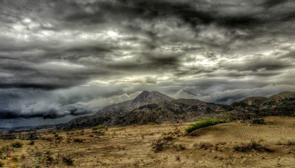 A mountain range under a dramatic cloudy sky with dark clouds and a barren landscape in the foreground. - Olive Oil Times