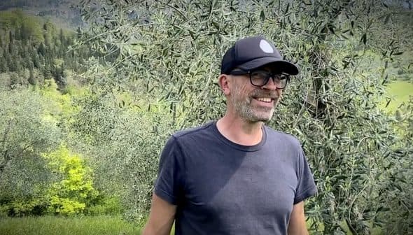 Man wearing a black cap and glasses smiling while standing in an olive grove. - Olive Oil Times